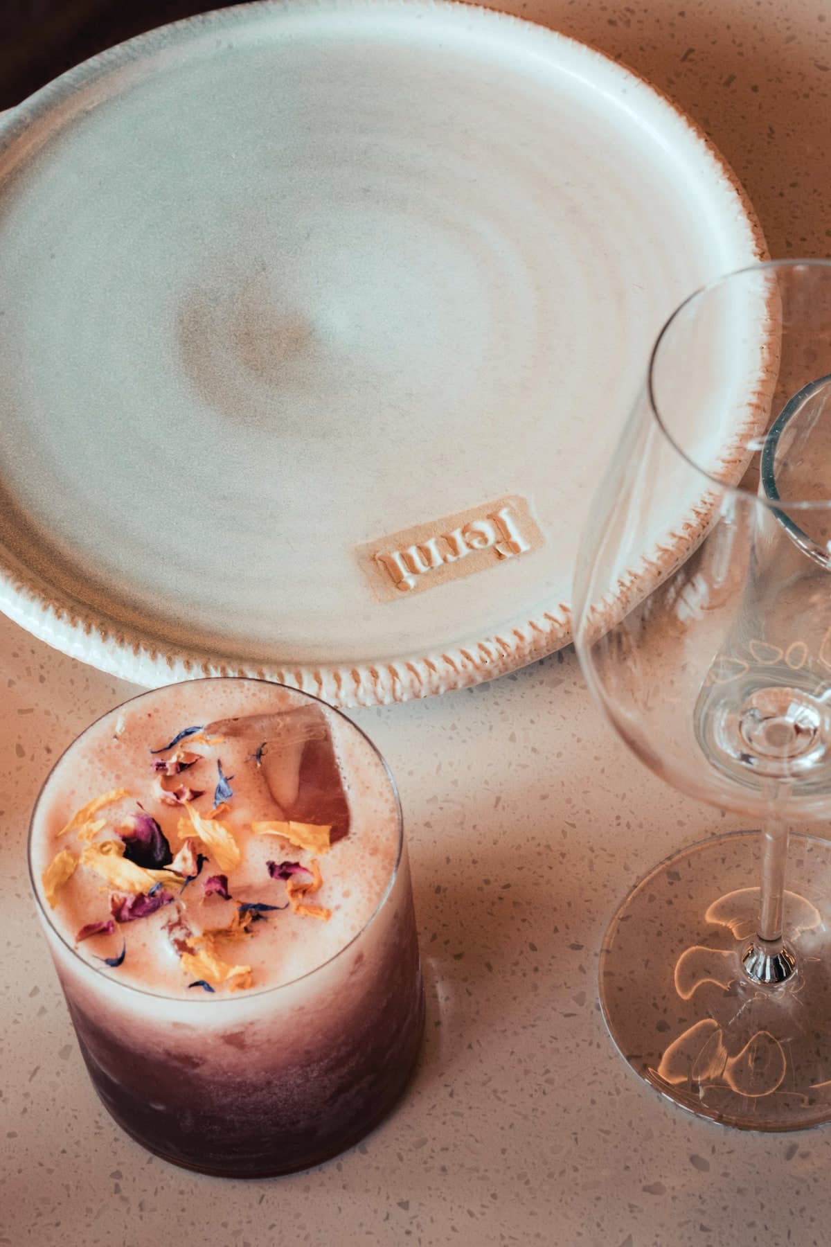 Cocktail sur glace aux pétales de fleurs, posé près d’une assiette de grès signée Ferni et d’un verre à vin