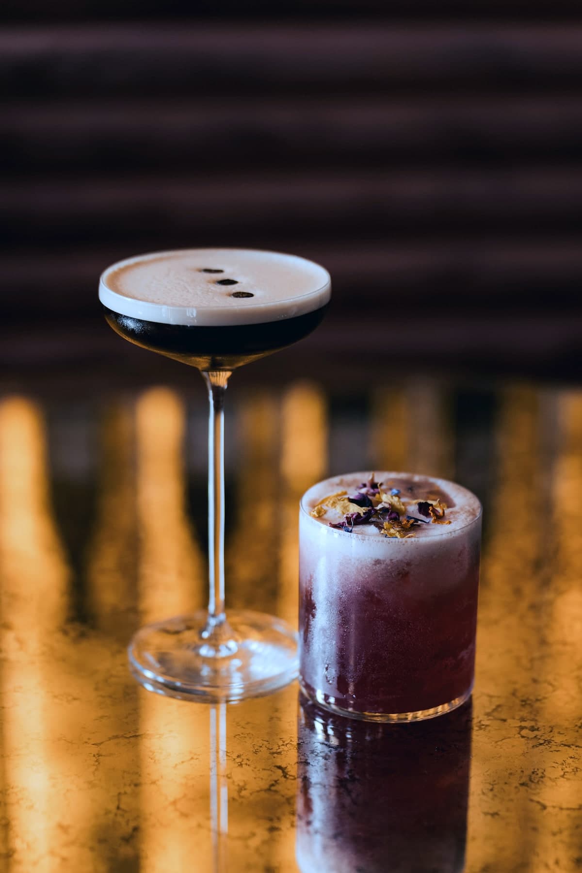 Deux cocktails sur un guéridon de laiton : un espresso martini en coupe et un sour violet aux pétales séchés