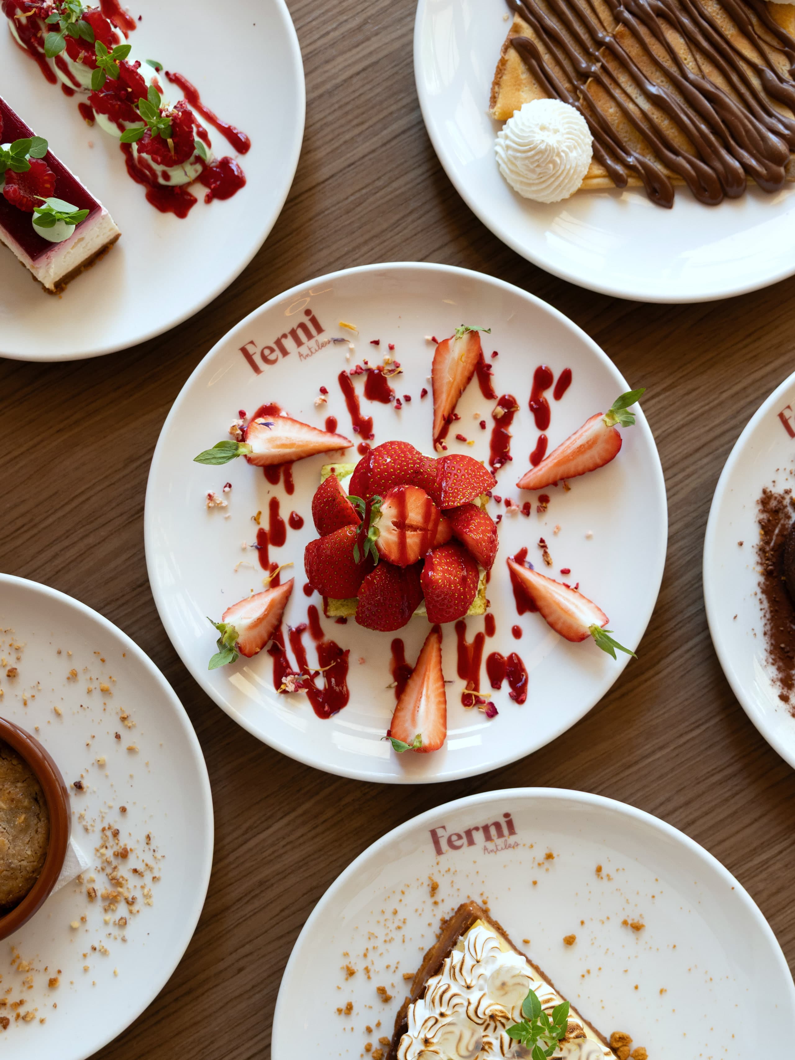Six assiettes de desserts vues de dessus sur une table de bois, dont un dressage de fraises au coulis sur assiette signée Ferni