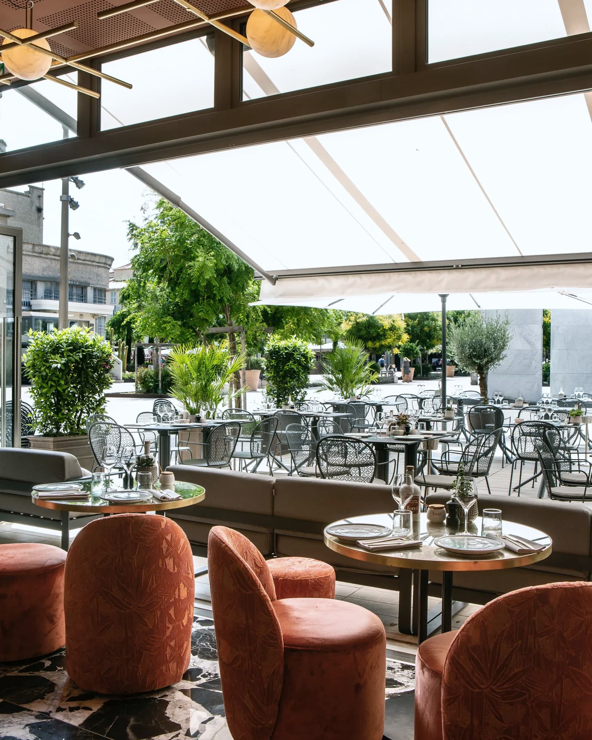 Terrasse sous la toile tendue, fauteuils terracotta et platanes de la place au second plan